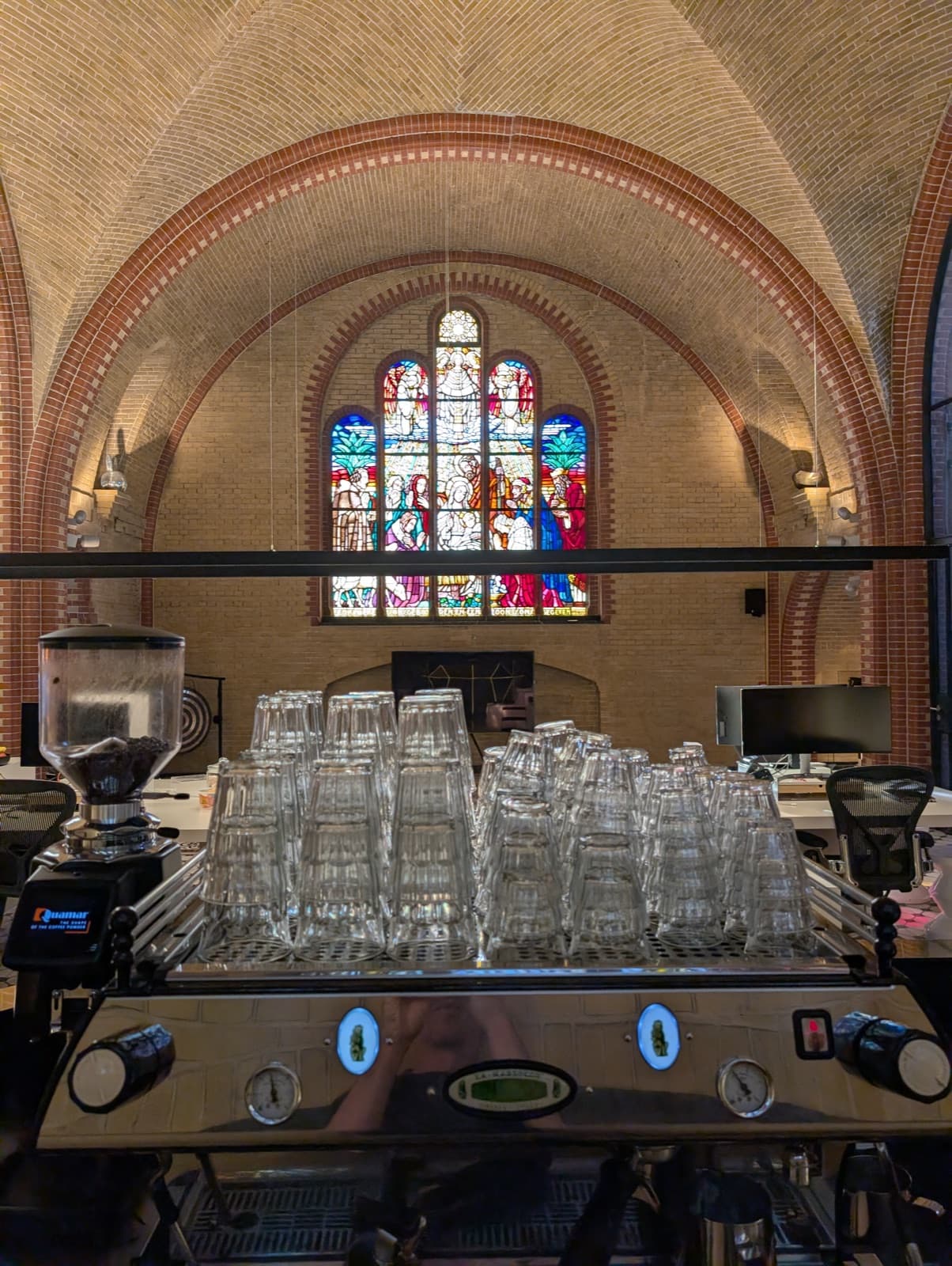 The vaulted brick ceiling of a former hospital chapel, a stained glass window above office desks and monitors, and an espresso machine stacked with glasses in the foreground