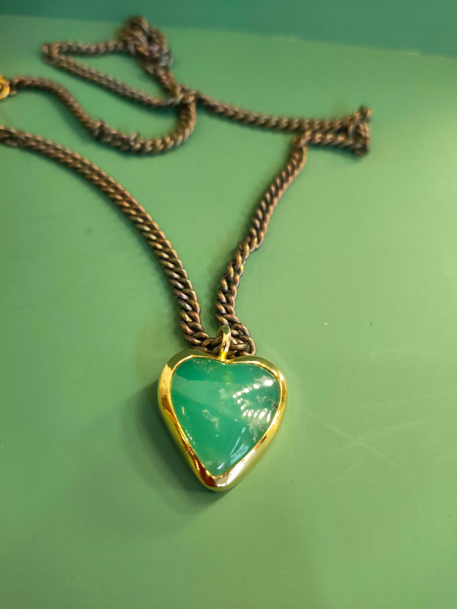 Original phone photo of a green heart pendant on a chain, on a green workbench mat